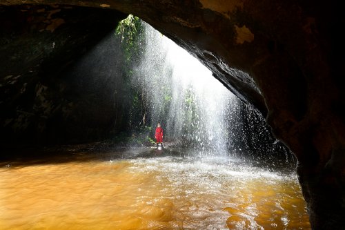 Caverna das Maos (Para, Brésil) - Cascade au soleil près de l'entrée de la grotte avec spéléo en fond et vasque d'eau jaune  en premier plan (horizontale)(SP-23-1248 )